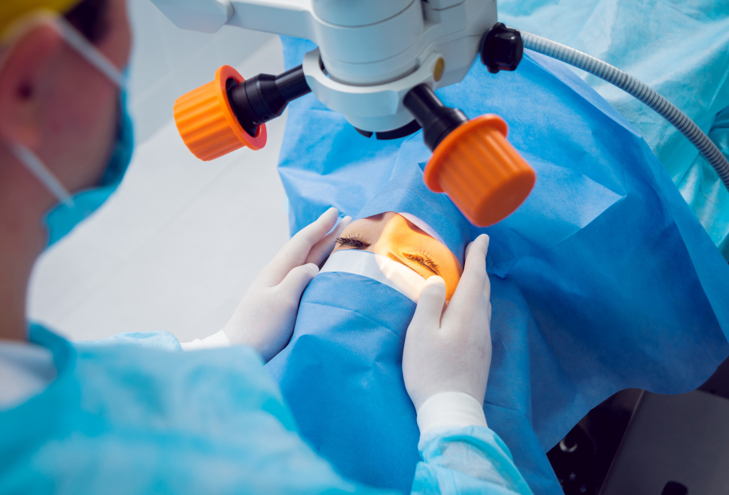 Surgeon in blue scrubs and mask performing eye surgery under a microscope, patient covered with sterile drapes.