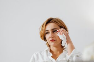 Woman wearing a white shirt dabbing her cheek with a tissue against a pale background, mid-face visible.