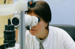 Person looking into a laboratory microscope, focusing through the eyepiece with a lab setting in the background, wearing a white coat and gray sweater.