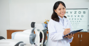 Female healthcare professional in a white coat smiles while writing on a clipboard in a clinic with an eye chart in the background.
