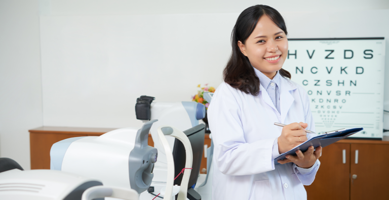 Female healthcare professional in a white coat smiles while writing on a clipboard in a clinic with an eye chart in the background.