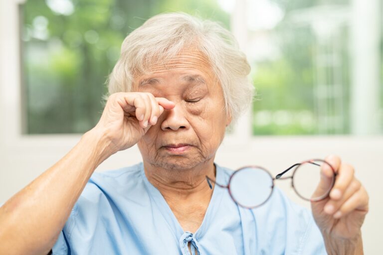 Asian senior woman with eyeglasses or vision glasses at home car – London Eye And Retina Specialist Centre – London Eye And Retina Specialist Centre Elderly woman in blue scrubs rubbing her eye while holding eyeglasses in one hand.