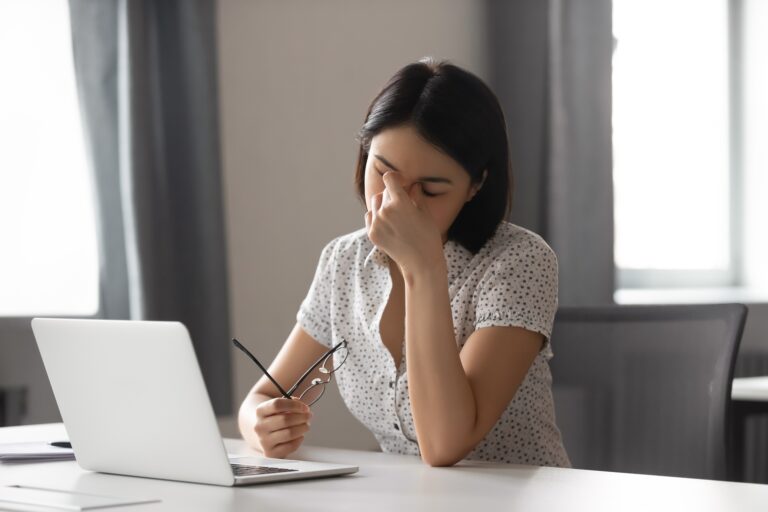 Woman at a desk with a laptop, rubbing her eyes and holding her glasses, looking tired or stressed.