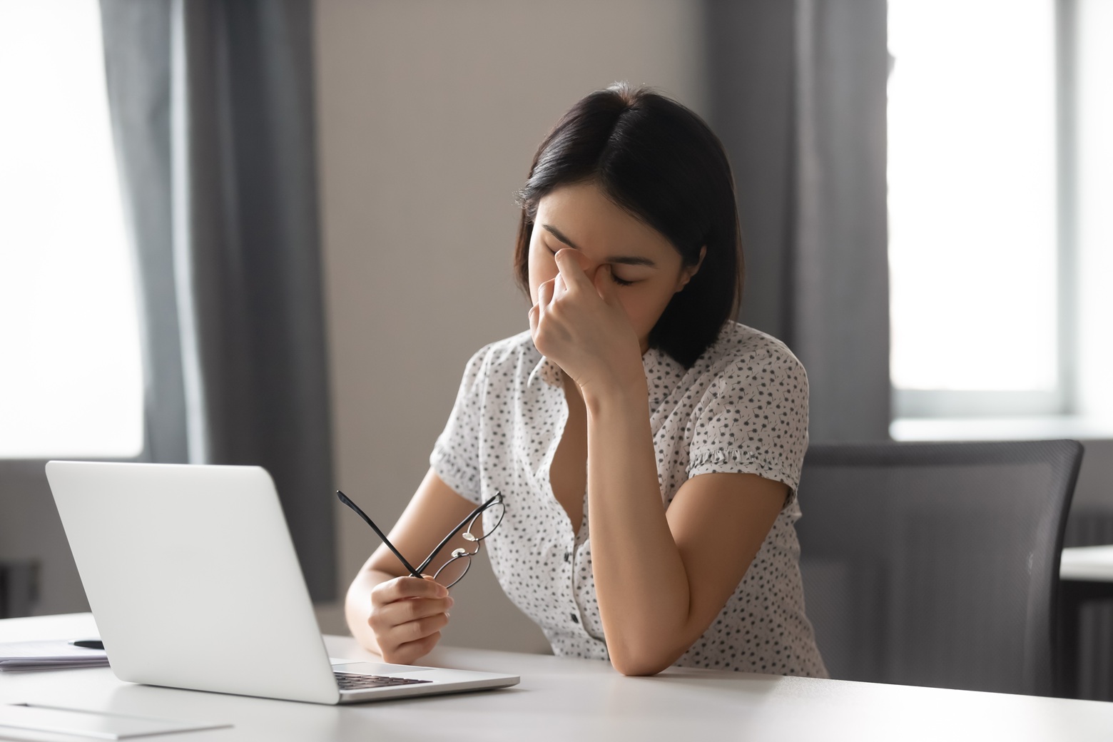 Woman at a desk with a laptop, rubbing her eyes and holding her glasses, looking tired or stressed.