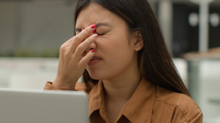 Overworked Asian woman girl working laptop in cafe suffer eyestrain tired chinese businesswoman freelancer overload with computer work rub eyes pain discomfort exhaustion blurry vision bad eyesight – London Eye And Retina Specialist Centre – London Eye And Retina Specialist Centre Woman with long dark hair rubbing her eyes with fingers while sitting at a desk, wearing a brown shirt.